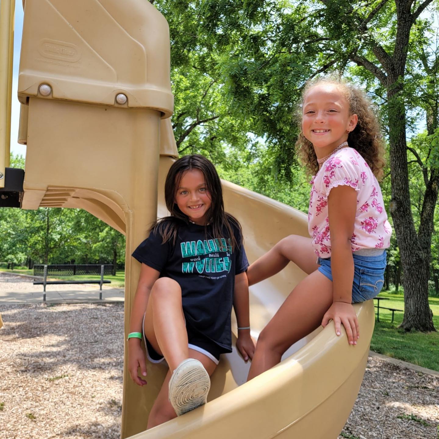 two girls sitting on a slide in the playground smiling for the camera